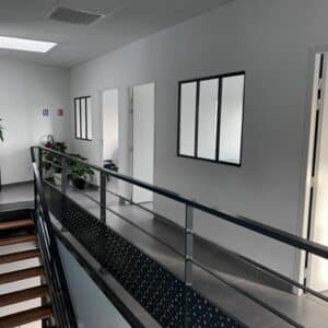 Couloir de bureau moderne avec escalier en bois, garde-corps métallique noir et plantes vertes décoratives.