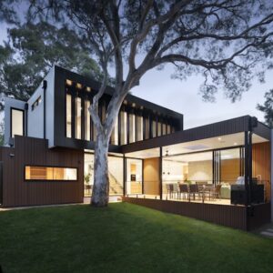 Maison contemporaine à deux étages, bardage bois et grandes baies vitrées avec arbre intégré au crépuscule.