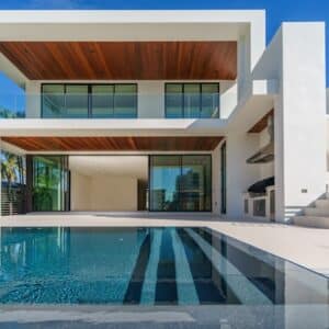 Villa moderne blanche avec piscine, plafonds en bois et grandes baies vitrées sous un ciel bleu dégagé.