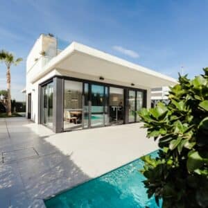 Villa moderne avec piscine, terrasse carrelée et grandes baies vitrées sous un ciel bleu ensoleillé.